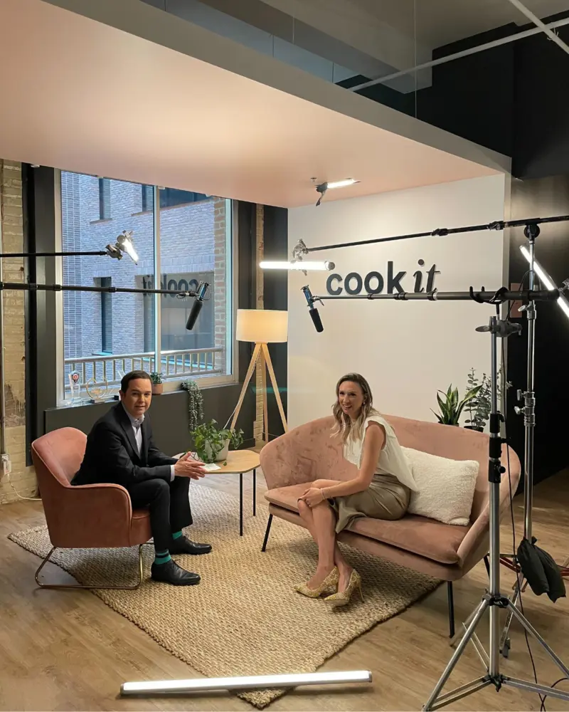 Judith Fetzer assise sur un canapé rose dans les bureaux de Cook it, en pleine entrevue filmée avec un homme en complet, entourés d’équipements de tournage