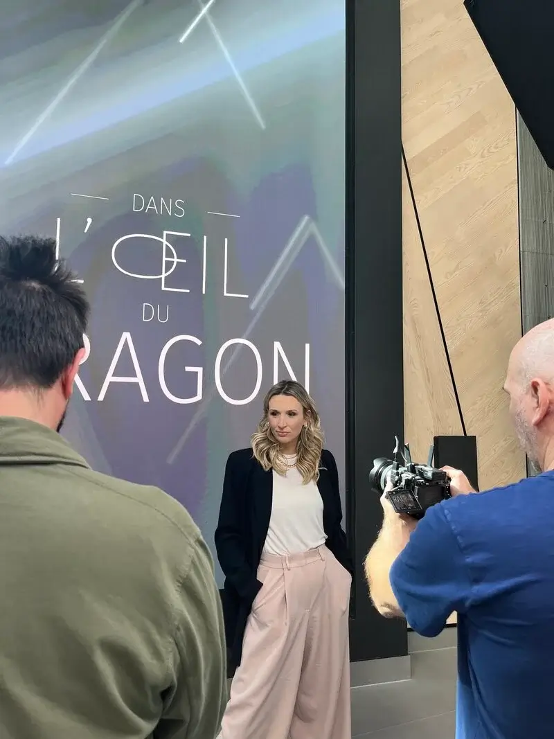 Judith Fetzer posant devant une affiche de l'émission “Dans l'œil du dragon”, habillée en pantalon pâle et veston foncé, lors d’un shooting photo en studio