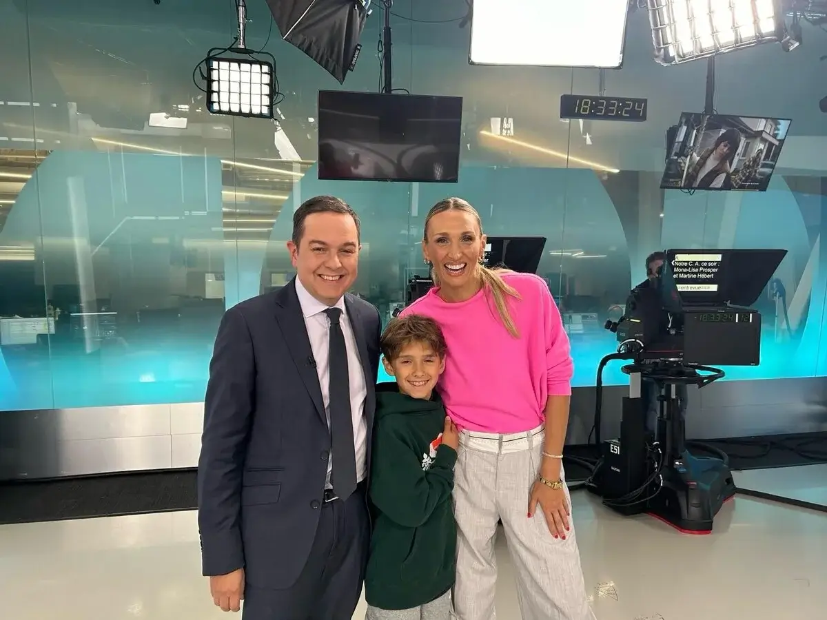 Judith Fetzer souriante en studio télé, vêtue d’un chandail rose, posant avec un journaliste en complet et un jeune garçon au milieu des caméras et éclairages