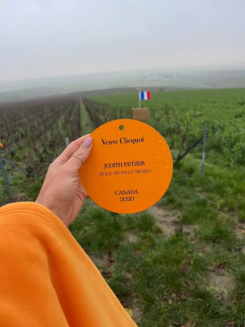Main tenant une plaque orange du Veuve Clicquot Bold Woman Award décernée à Judith Fetzer en 2020, dans un vignoble brumeux en France avec un drapeau tricolore au loin