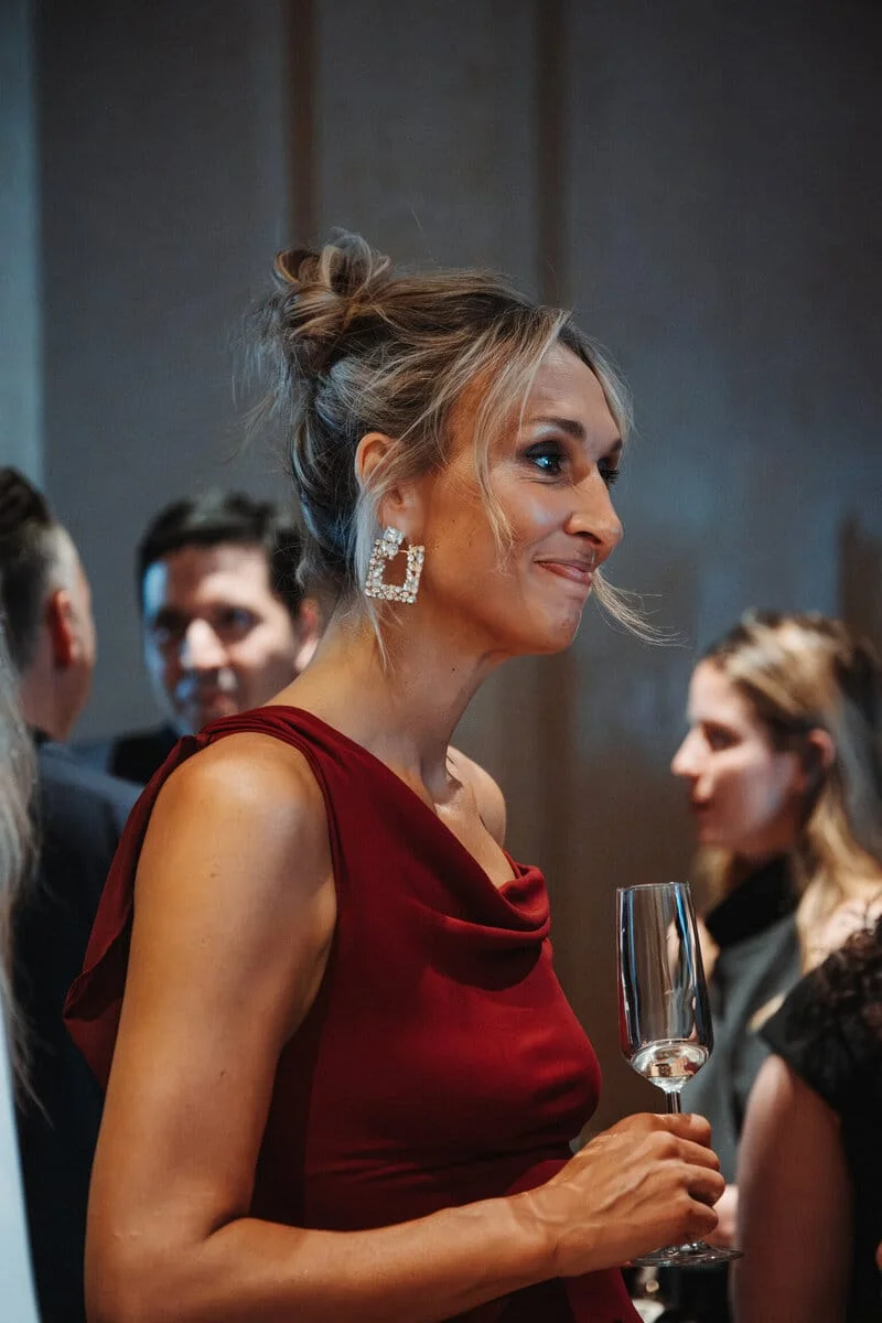 Judith Fetzer élégamment vêtue d’une robe rouge, tenant une flûte à champagne et souriant lors d’un événement mondain